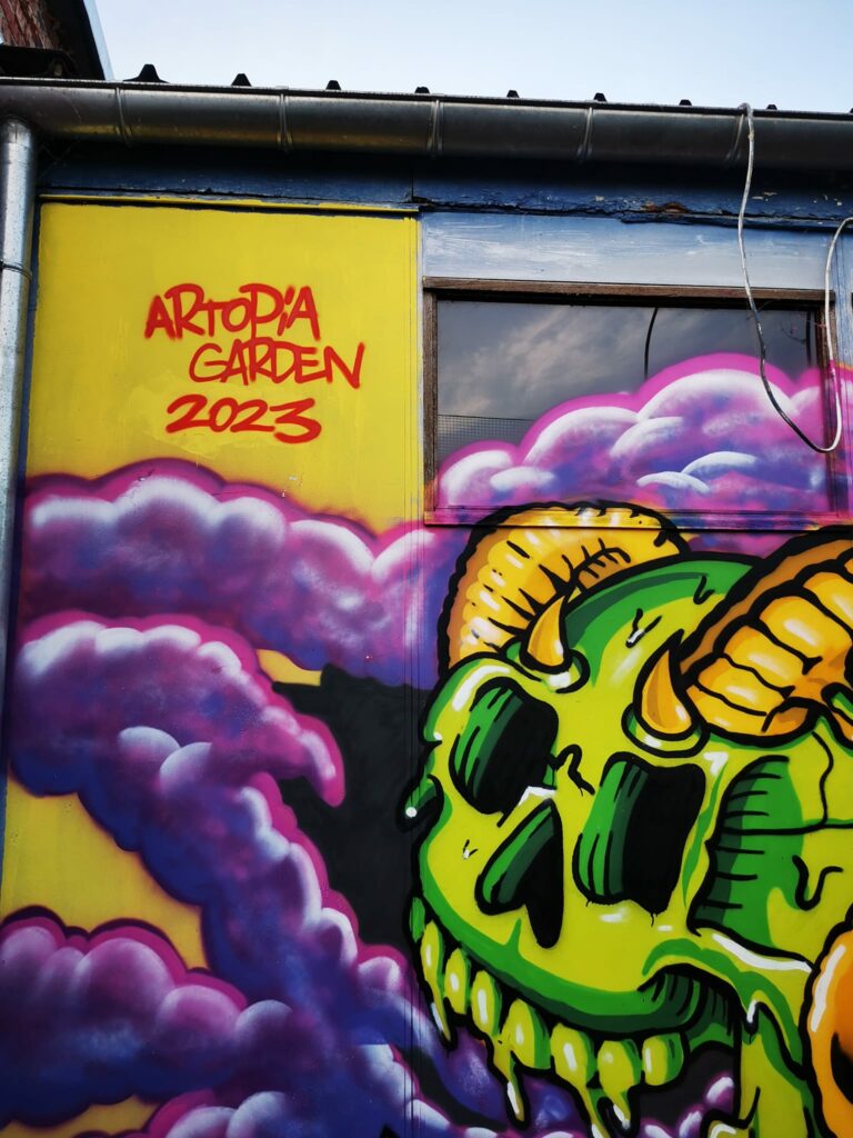 Un graffiti représentant un crâne vert entouré de nuages violets avec « Artopia Garden 2023 » en écriture rouge